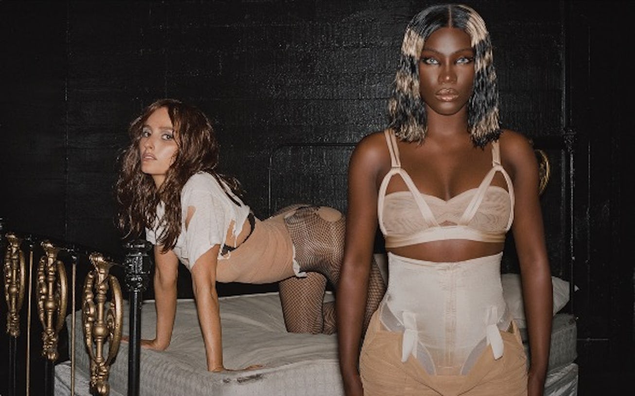 Two models pose on a bed with a dark backdrop. One is on all fours wearing sheer clothing, while the...