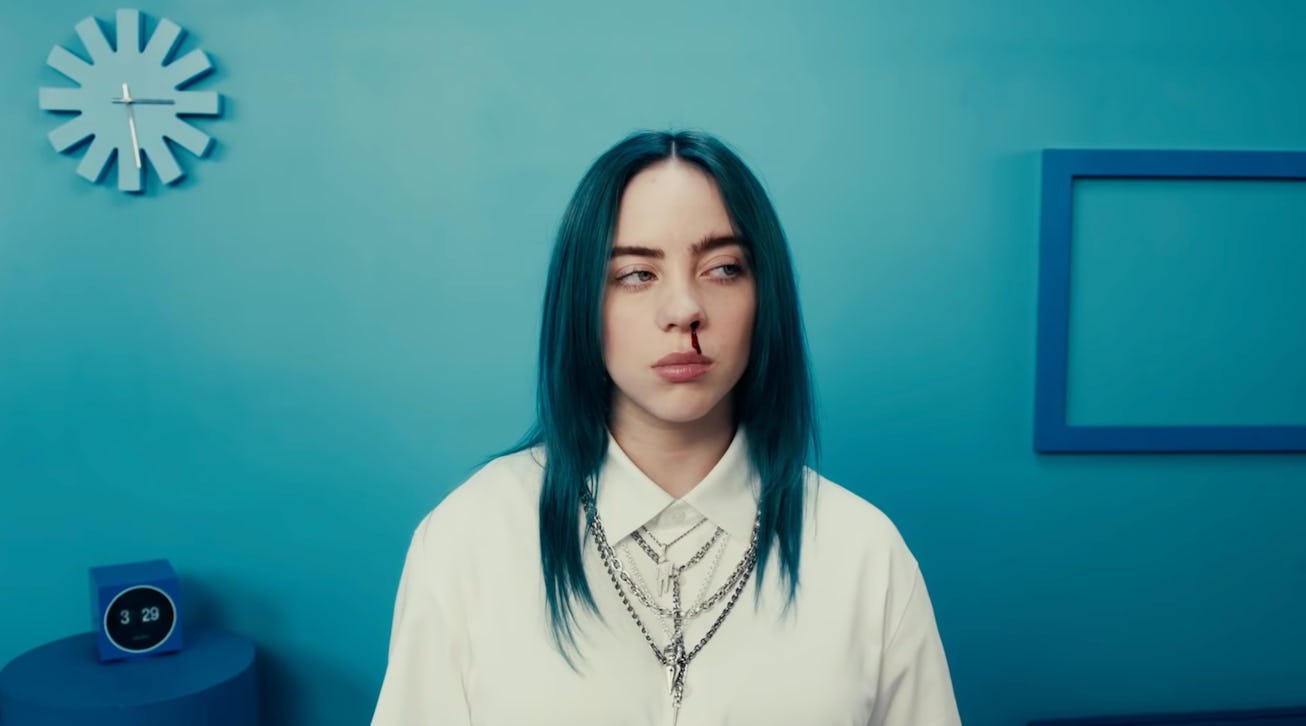 Woman with blue hair and a white shirt standing in a blue room, looking slightly to the side with a ...