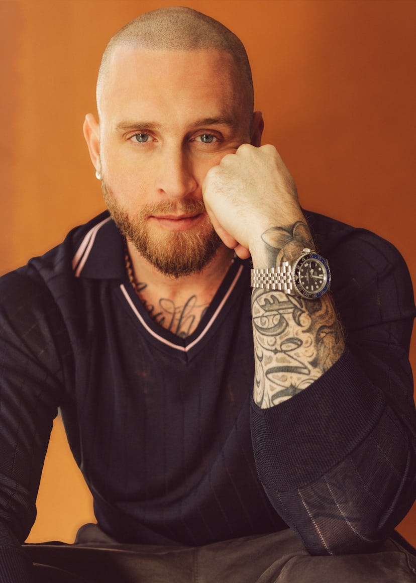 A man with a shaved head and a beard rests his chin on his hand, wearing a dark sweater. He has visi...